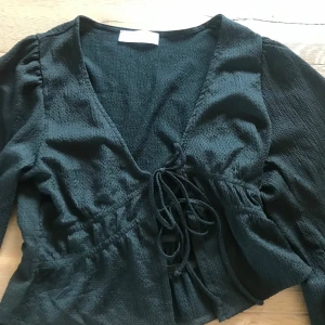 Svart omlottopp med snörning - En svart omlottopp med v-ringning och långa ärmar. Toppen har snörning framtill i midjan och är croppad med lätt puffade ärmar. Materialet är strukturerat och känns mjukt mot huden. Perfekt för dig som gillar trendiga detaljer och en snygg siluett.