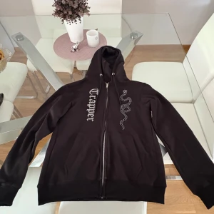 Svart Rhinestone hoodie med dragkedja - Svart hoodie med huva, hel dragkedja och snyggt tryck framtill – ena sidan har texten 'Trapper' i gotisk stil och andra sidan en glittrig orm. Perfekt för dig som gillar streetwear och vill sticka ut lite extra. Inga fel på den utan säljer pga för liten.