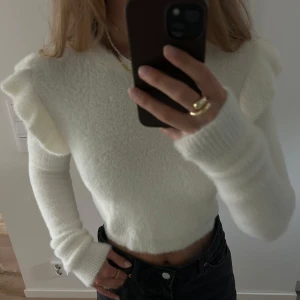 Vit fluffig croppad stickad tröja - Supermjuk och fluffig vit stickad tröja med croppad passform och volangdetaljer på axlarna. Långärmad modell (något längre ärmar än ”vanligt”) med rund halsringning 🤩🥰