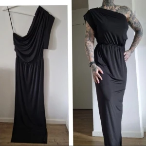 Svart långklänning oneshoulder - Svart långklänning med oneshoulder-design och draperad överdel. Klänningen har resår i midjan och är tillverkad i ett mjukt, stretchigt material som faller snyggt längs kroppen. Perfekt för dig som gillar en stilren och edgy look.