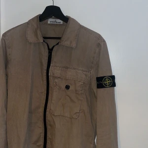 overshirt från Stone Island - Snygg beige overshirt från Stone Island med klassisk krage, bröstficka med knapp och ikonisk patch på ärmen. Jackan har dragkedja framtill och är tillverkad i bomull, perfekt för lager-på-lager-stil. Passar dig som gillar clean och stilren look. Den är i storlek small och sitter perfekt på dig som vill vara stilren till vardagen. Köpt för något år sedan så kvittot har försvunnigt i flytt. 