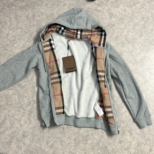 Grå hoodiejacka från Burberry - Snygg grå hoodiejacka från Burberry med klassiskt rutigt foder i huvan. Jackan har dragkedja framtill, fickor och ribbade muddar. Tillverkad i mjuk bomull med en skön känsla. Perfekt för dig som vill ha en stilren look med en touch av lyx.