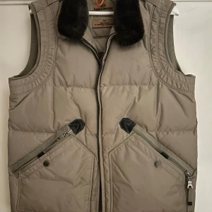 Beige dunväst från Parajumpers - Snygg beige dunväst från Parajumpers med mörkbrun fuskpäls vid kragen och coola detaljer som stora snedställda fickor med dragkedja. Västen har klassisk PJS-logga på ryggen och robust känsla, perfekt för lager-på-lager-stil. Är storlek L men sitter bäst för något med M i vanliga fall. 