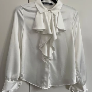 Vit blus med volang från Zara - Snygg vit blus från Zara i glansigt material med stora volanger framtill och vid ärmslut. Klassisk krage med knappdetalj och långa ärmar. Perfekt för dig som vill sticka ut med en elegant och trendig look.
