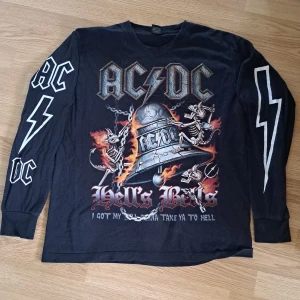 AC/DC Hell's Bells långärmad tröja - Svart långärmad AC/DC-tröja från Rockvolution med stort Hell's Bells-tryck fram och bak. Skelett, eld och blixtar ger en riktigt rockig vibe. AC/DC-logga och blixtar på ärmarna. Perfekt för dig som älskar bandmerch och vill sticka ut.