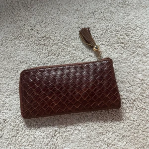 Brun flätad plånbok i skinn - Snygg brun plånbok i flätat skinn med guldfärgad dragkedja och tofsdetalj. Insidan har flera kortfack och ett separat myntfack med dragkedja. Perfekt för dig som vill ha en stilren och praktisk accessoar.