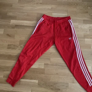 Röda Adidas mjukisbyxor med vita ränder - Snygga röda mjukisbyxor från Adidas med klassiska vita ränder längs sidorna och broderad logga på låret. Byxorna har snörning i midjan och muddar vid bensluten. Perfekta för chill eller träning.