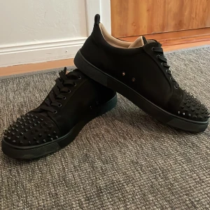 Svarta sneakers med nitar från Louboutin - Svarta sneakers i skinn med coola svarta nitar över tån och klassisk röd sula. Skorna har snörning och en stilren, chunky look som sticker ut. Perfekta för dig som vill ha något edgy och exklusivt i garderoben.