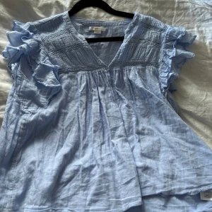 Ljusblå blus med volang från Gina Tricot - Supersöt ljusblå blus från Gina Tricot med v-ringning och volangdetaljer på axlarna. Blusen har korta ärmar och är tillverkad i ett luftigt bomullstyg, perfekt för varmare dagar. Smockad upptill och lös passform för en avslappnad look.