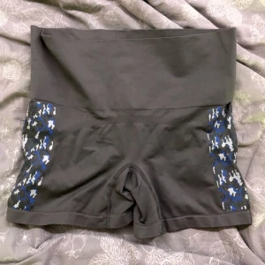 Grå träningstights-shorts från Crivit - Grå höga träningstights-shorts från Crivit med bred midja och coola blå, vita och svarta camouflage-detaljer på sidorna. Tillverkade i stretchigt material med polyester och elastan för maximal komfort under träning. Perfekta för gymmet eller löprundan.