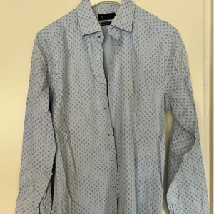 Ljusblå mönstrad skjorta från Riley - Snygg ljusblå skjorta från Riley i slim fit-modell. Skjortan har ett diskret geometriskt mönster i mörkblått, klassisk krage och knäppning framtill. Perfekt för dig som gillar stilrena detaljer och en smalare passform. Köptes på Brother för 1100kr