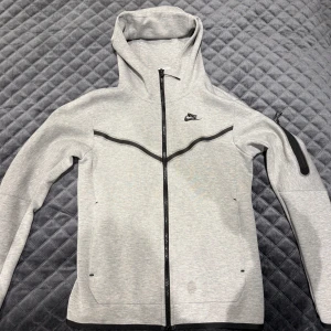 Grå Nike tech - Snygg grå vindjacka från Nike i storlek XS. Jackan har hel dragkedja, huva och svarta detaljer längs bröstet och ärmen. Perfekt för dig som gillar sportig stil och vill ha något lätt och bekvämt. Klassisk Nike-logga på bröstet. OBS den har en liten fläck.