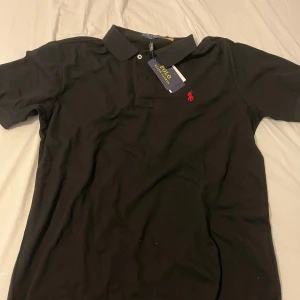 Ralph lauren pike tröja  - Klassisk svart pikétröja från Polo Ralph Lauren med röd broderad logga på bröstet. Tröjan har krage, korta ärmar och knäppning framtill. Slim fit-modell i mjuk bomull som ger en clean och stilren look.