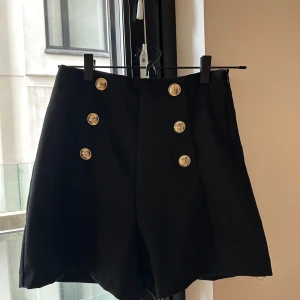 Svarta shorts med guldknappar från Zara - Stilrena svarta shorts från Zara med hög midja och sex snygga guldknappar framtill. Klassisk och lite dressad look som passar perfekt till en trendig outfit. Materialet känns mjukt och har en lätt struktur. Perfekta för dig som gillar en chic vibe.