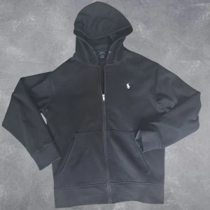 Polo Ralph Lauren zip hoodie - Svart hoodie från Polo Ralph Lauren med dragkedja framtill och klassisk vit logga på bröstet. Tröjan har huva med snörning och två stora fickor framtill. Perfekt för en chill och stilren look. Storlek L men passar även M.