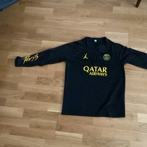 Svart PSG långärmad fotbollströja - Snygg svart långärmad PSG-tröja från Jordan med gula detaljer och Qatar Airways-tryck på bröstet. Paris-text på ärmen och klubbmärke på bröstet. Perfekt för dig som vill sticka ut på planen eller på läktaren.Rekommenderar inte för personer som är 178+ cm, helt ny aldrig använt 