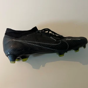 Nike Mercurial Air Zoom fotbollsskor - Svarta Nike Mercurial Air Zoom fotbollsskor med snörning och låg profil. Ovandel i syntetmaterial med diskret mönster och silverdetaljer. Sulan har gula och svarta inslag samt fasta dobbar för bra grepp på planen. Perfekta för dig som vill ha speed och kontroll.