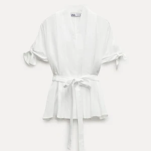 Vit omlottblus med knyt från Zara XS - Superfin vit omlottblus från Zara i luftig bomull/linne-blandning. Korta ärmar med knyt, v-ringning och bälte i midjan för snygg passform. Perfekt till jeans eller kjol när du vill ha en clean och fräsch look.