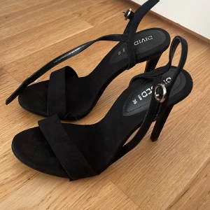 Svarta klackade sandaler H&M Divided - Svarta sandaler med hög klack från H&M Divided. Snygg minimalistisk design med bred rem över tårna och smal rem runt vristen med silverfärgat spänne. Gjorda i syntetmaterial med öppen tå och platt sula framtill. Perfekta för dig som gillar stilrena vibes.