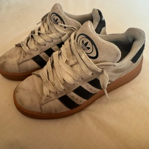 Adidas sneakers med svart-vita ränder - Klassiska Adidas sneakers i beige mocka med svarta ränder och gummisula i brunt. Skorna har snörning och den ikoniska Adidas-loggan på plösen. Perfekta för dig som gillar retrostil och streetwear.