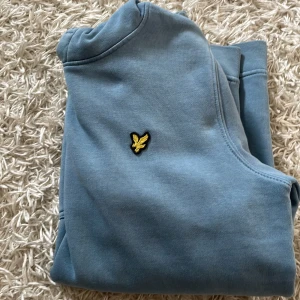 Blå hoodie från Lyle & Scott - Snygg blå hoodie från Lyle & Scott med klassisk gul logga på bröstet. Tröjan har huva med snörning, känguruficka och långa ärmar. Perfekt för dig som gillar en enkel men stilren look. Tillverkad i mjuk bomull för skön känsla. Passar på 10/13 åringar 