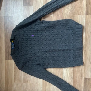 Grå kabelstickad tröja Polo Ralph Lauren - Mörkgrå kabelstickad tröja från Polo Ralph Lauren med lila broderad logga på bröstet. Tröjan har rund halsringning, långa ärmar och ribbade muddar. Perfekt för dig som gillar klassisk och stilren look.