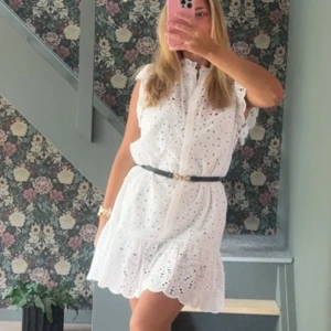 Vit kort klänning med broderade detaljer - Superfin vit kort klänning med broderade hål och volangkant. Klänningen har ärmlös design, knappar framtill och ett smalt bälte i midjan som ger en snygg siluett. Perfekt för varma dagar och sommarhäng. Stolek M men passar S också, satte skärp runt 