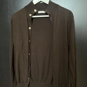 Brun kofta med knappar från SuitSupply  - Mörkbrun kofta från SuitSupply med långa ärmar och ribbad krage. Klassisk modell med knäppning framtill och mjukt, tunt material som känns skönt mot huden. Perfekt att slänga över en t-shirt eller linne för en chill look. (Litet hål på ena ärmen, knappt synbart). Krymt något därav säger jag STL S