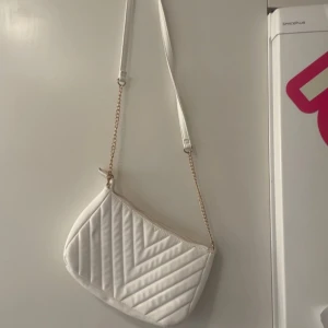 Vit quiltad axelväska - Snygg vit axelväska med quiltat mönster och dragkedja. Väskan har en kedja som axelrem vilket ger en stilren look. Perfekt för att bära med sig det viktigaste.
