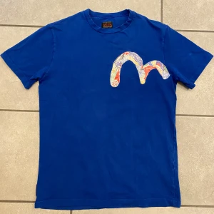 Evisu vintage t-shirt  - Säljer en blå vintage designer t-shirt från Evisu med ett färgglatt mönster på bröstet. T-shirten har en rund hals och korta ärmar. Perfekt för en avslappnad stil. Provad några gånger men den är som ny. Längd 68cm samt Bredd 47cm.