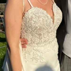 Vit bröllopsklänning med smala axelband och låg rygg - Elegant vit klänning med spetsdetaljer och smala axelband. Klänningen har en vacker öppen rygg och knappar längs ryggen. "Formfitting" med plats att sy ut lite. Köptes för 14.000kr his Dream design i Motala. Finns en överkjol som jag ej använt som medkommer