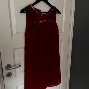 Röd ärmlös klänning från Vero Moda - Snygg röd ärmlös klänning från Vero Moda med glittriga detaljer runt halsen och axlarna. Perfekt för att sticka ut med sin eleganta design och fina detaljer. Klänningen har en luftig passform och är gjord i ett lätt material.