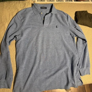 Blå pikétröja från Ralph Lauren - Säljer en klassisk blå pikétröja från Ralph Lauren med lång ärm och krage. Tröjan har knappar framtill och den ikoniska loggan broderad på bröstet. Perfekt för en stilren look. Storlek medium. Fläckfri.