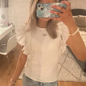 Vit topp med volangdetaljer - Säljer en söt vit topp med volangdetaljer på axlarna. Toppen har en tight passform och är perfekt för en stilren look. Har en sminkrand men som inte snyns när man bär plagget, går antagligen bort i tvätten💕