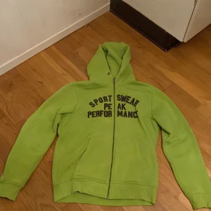 Grön huvtröja från Peak Performance - Säljer en grön huvtröja från Peak Performance med texten 'SPORTSWEAR PEAK PERFORMANCE' på framsidan. Tröjan har dragkedja och är tillverkad av 80% bomull och 20% polyester. Perfekt för en avslappnad stil.