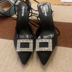 Svarta klackskor med strassdetalj - Eleganta svarta klackskor från Styleloop med spetsig tå och glittrande strassdetalj på ovansidan. Skorna har en hög klack och knytband som ger en stilren look.