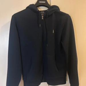 Mörkblå hoodie från Polo Ralph Lauren - Säljer en stilren mörkblå  hoodie från Polo Ralph Lauren. Den har dragkedja framtill och en klassisk huva. Perfekt för en avslappnad look. 9/10 skick. 