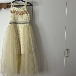 Beige spetsklänning från Miss Liwi - Säljer en elegant beige klänning från Miss Liwi med spetsdetaljer och volang. Klänningen har en vacker öppen rygg och dragkedja baktill. Den är ärmlös och har en lång, flödande kjol i tyll. Perfekt för speciella tillfällen! Använt 1 gång, 1 diamant har fallit. Köpte den för 2000kr men jag säljer den för 1000kr priset kan diskuteras. Klänning för 11-12 år. (festklänning)