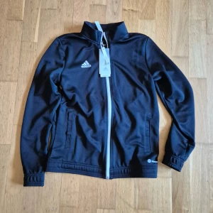Svart tracktop från Adidas - Säljer en svart tracktop från Adidas med dragkedja och två fickor framtill. Jackan har en stilren design med Adidas-logga på bröstet. Perfekt för träning eller en sportig look.