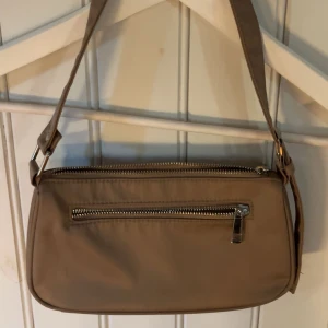 Beige axelväska från Bik Bok - Snygg beige axelväska från Bik Bok med praktisk dragkedja framtill. Perfekt för att bära dina nödvändigheter med stil. Väskan har en justerbar axelrem och är tillverkad i ett slitstarkt material.