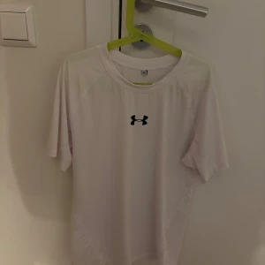 Compression shirt från Under Armour - Säljer en vit compression shirt från Under Armour med korta ärmar och logotyp på bröstet. Perfekt för träning. Skön och lätt att bära.