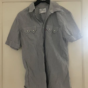 Randig kortärmad skjorta från Levi's - Snygg kortärmad skjorta från Levi's i blått och vitt randigt mönster. Skjortan har knappar framtill och två bröstfickor med knappdetaljer. Perfekt för en avslappnad stil.