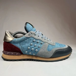 Valentino Rockrunner sneakers i jeans och mocka - Snygga Valentino Rockrunner sneakers i en cool mix av jeans och mocka. Skorna har en unik design med grå snörning och detaljer i svart och vinrött. Den guldiga hälkappan och de ikoniska nitarna på sidan ger en extra edge. Strl 42 men passar mer 43. Nypris 6000. 