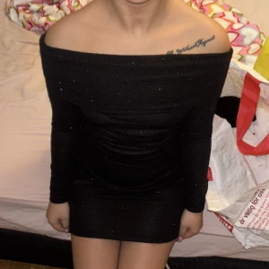 Svart glittrig klänning - Säljer en elegant svart glittrig klänning med offshoulder-design. Klänningen är kort och har långa ärmar, perfekt för en kväll ute. Den glittriga ytan ger en extra touch av glamour. Passar bra för festliga tillfällen.