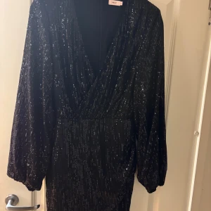 Svart glittrig klänning från Nelly - Snygg svart klänning från Nelly med glittriga detaljer. Den har en v-ringad design och långa ärmar, perfekt för en festkväll. Klänningen har en elegant passform som framhäver figuren.