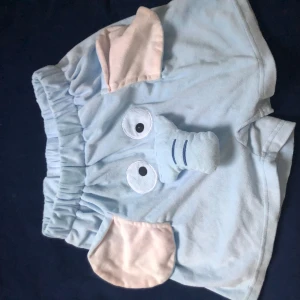 Blå shorts med elefantdesign -  ljusblå shorts med elefantmotiv, inklusive öron och snabel. Elastisk midja för bekväm passform. Snabeln har en pipleksak 