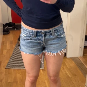 Blå low Waits jeansshorts från pull&bear - Säljer ett par snygga blå jeansshorts med fransiga kanter och slitna detaljer. Shortsen har en klassisk femficksdesign och knappgylf. Perfekta för en avslappnad stil.