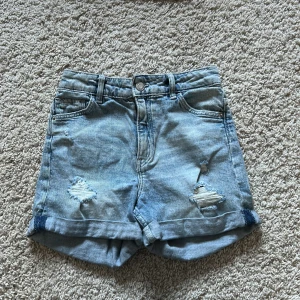 Blå jeansshorts med slitningar - Säljer ett par coola blå jeansshorts från H&M med slitningar framtill. De har en avslappnad passform och är midwaist