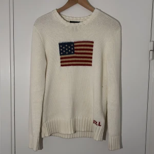 Vit stickad tröja från Ralph Lauren - Säljer en vit stickad tröja från Ralph Lauren med en broderad amerikansk flagga på bröstet. Tröjan har långa ärmar och ribbstickade muddar. Perfekt för en stilren och klassisk look.
