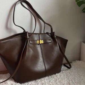 Brun axelväska från Gina Tricot - Snygg och rymlig brun axelväska från Gina Tricot i syntetmaterial. Väskan har ett stilrent spänne i guld och långa axelremmar för bekväm bärning. Perfekt för att bära med sig allt man behöver under dagen. Endast använd 1 gång, den är mörkbrun! 